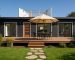 Casa modular moderna con revestimiento oscuro, una gran terraza de madera con muebles de exterior y una puerta de entrada de madera.
